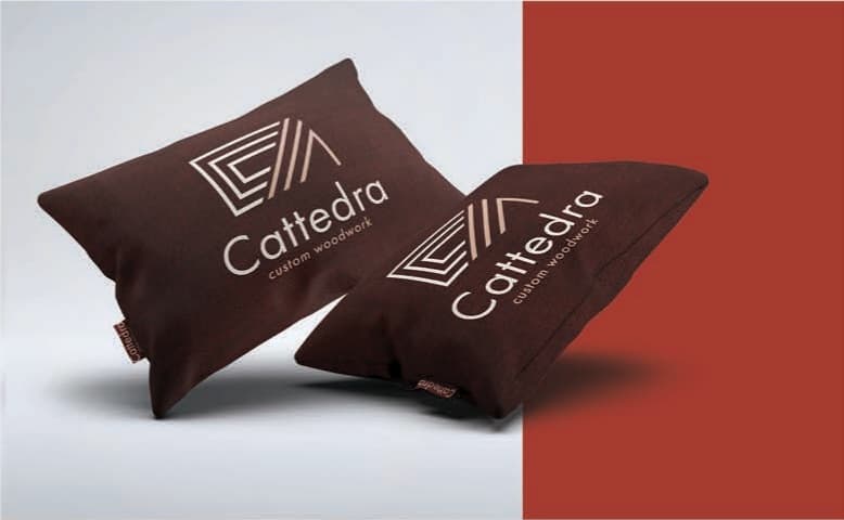 Almofadas com bordado do monograma Cattedra em tons de vinho e areia, conforme a aplicação original do brandbook.